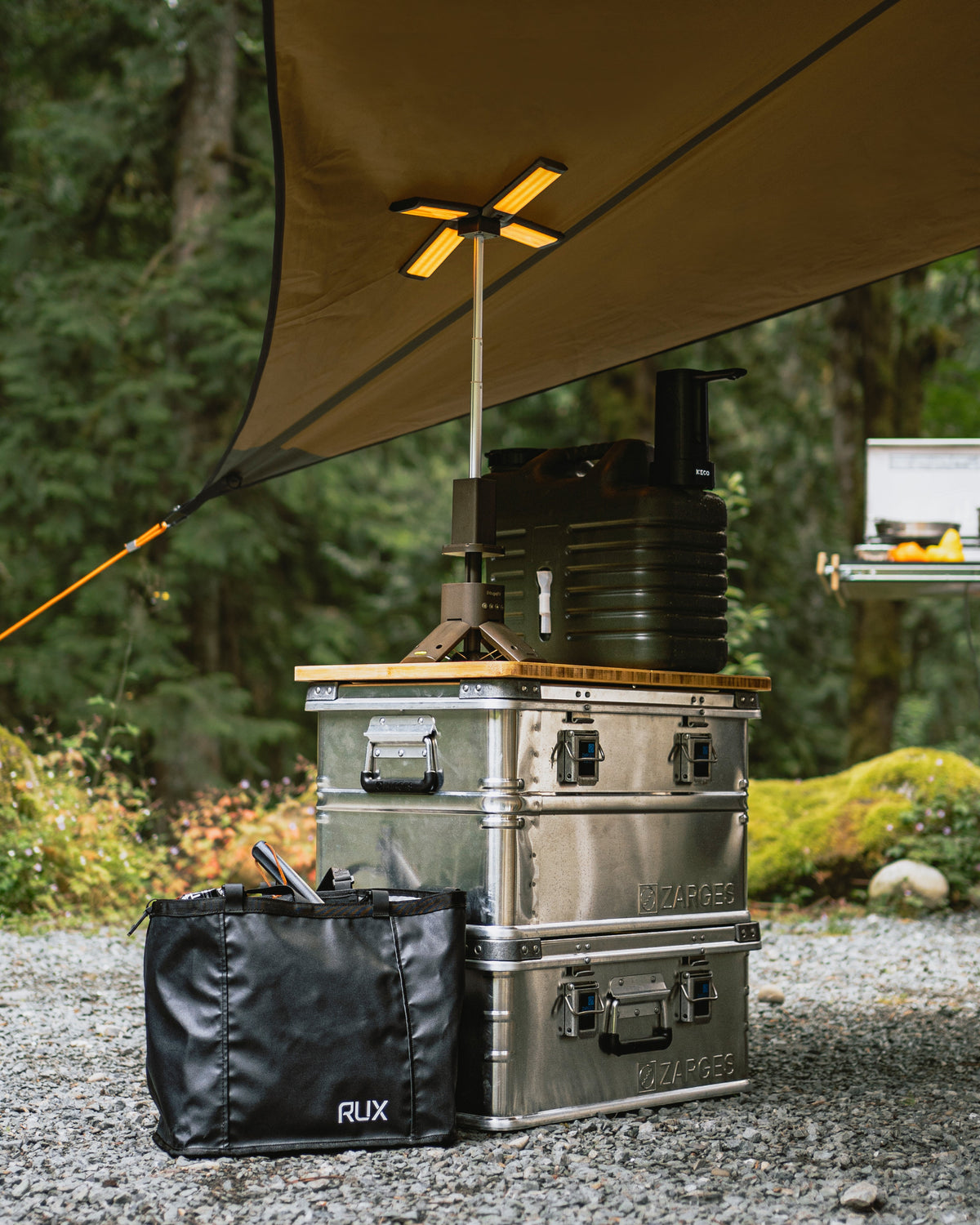 Stacked ZARGES K470-40678 cases with a wood top holding a water jug and lantern under a forest camp tarp