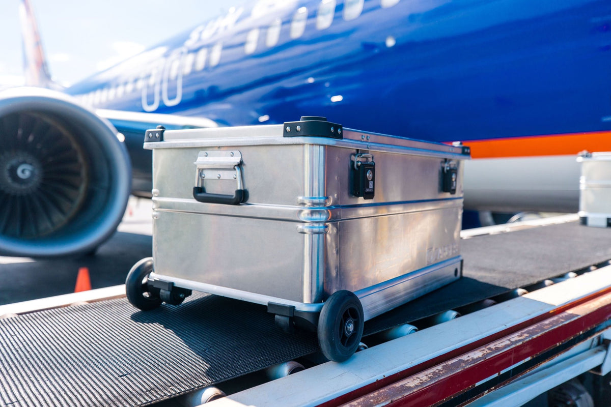 ZARGES K424 XC-41813 case riding a baggage belt loader beside a blue airliner on the tarmac