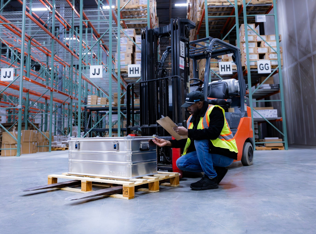 Worker kneeling with a clipboard beside a ZARGES K470-40567 aluminum case on a wooden pallet