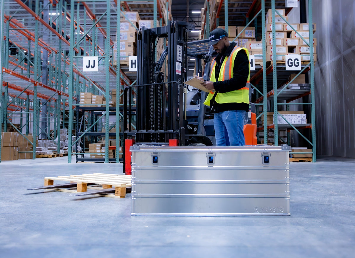 ZARGES K470-40580 case on a warehouse floor as a worker in a safety vest checks a clipboard by a forklift