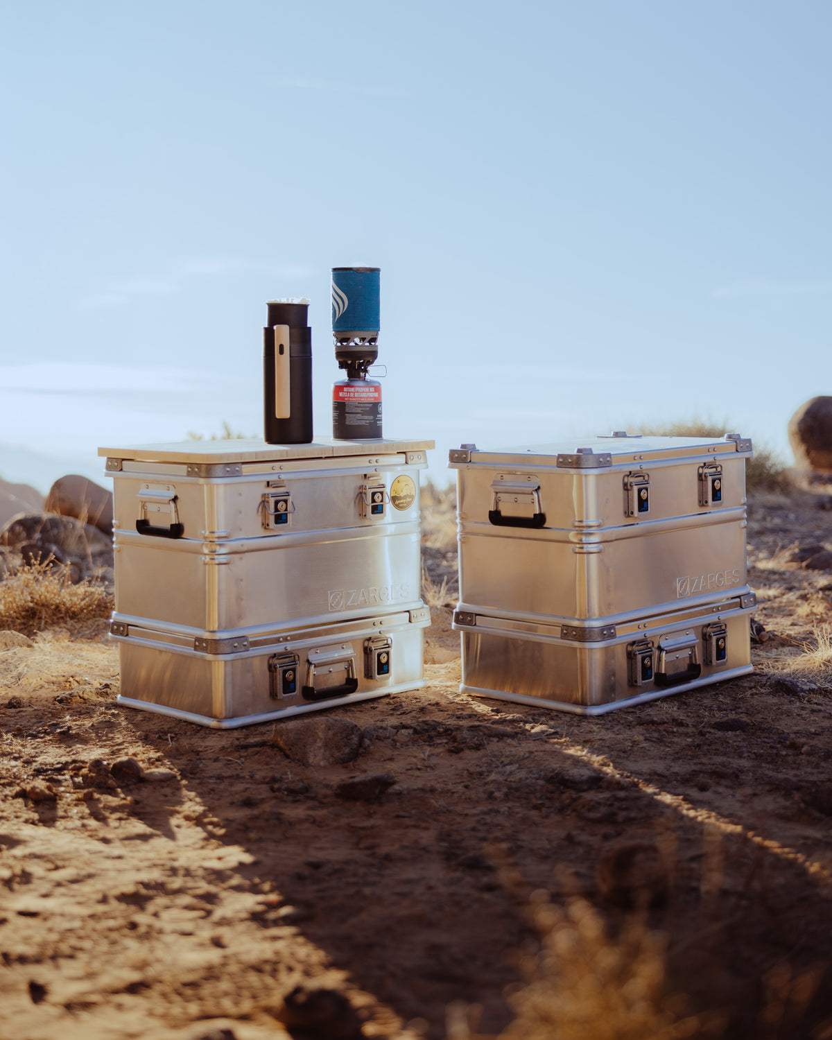 Two stacks of ZARGES K470-40678 cases on desert ground at sunset, a camp stove and bottle on top