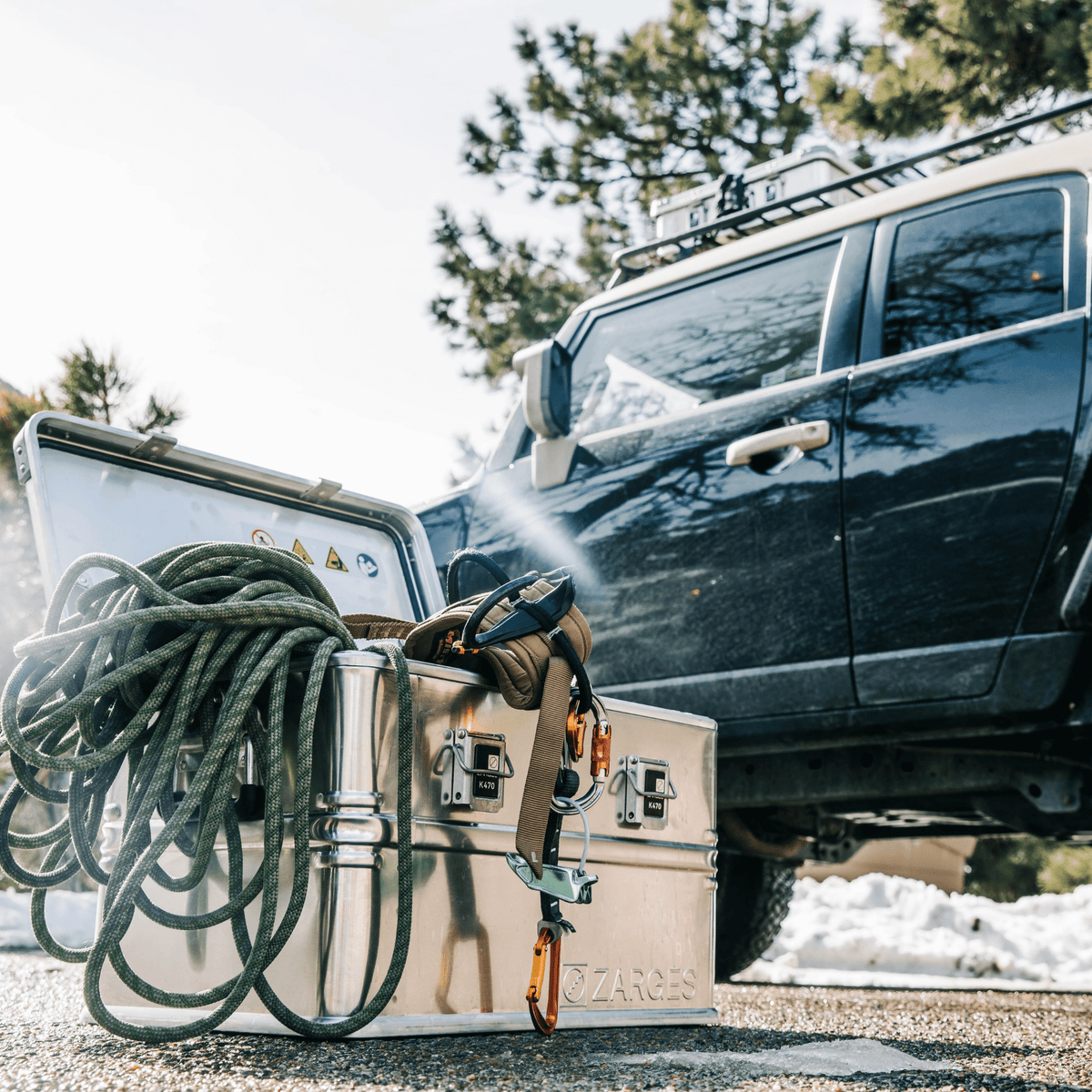 Open ZARGES K470-40678 case loaded with climbing rope and carabiners beside a dark SUV in the snow