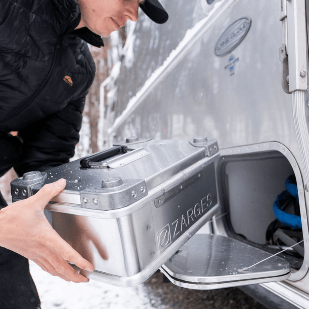Person sliding a ZARGES K470-40835 aluminum case into a storage compartment on a silver camper