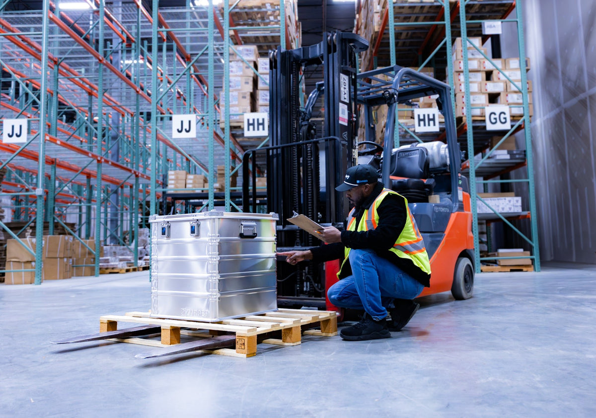 Warehouse worker kneeling with a clipboard beside a ZARGES K470-40836 case on a pallet