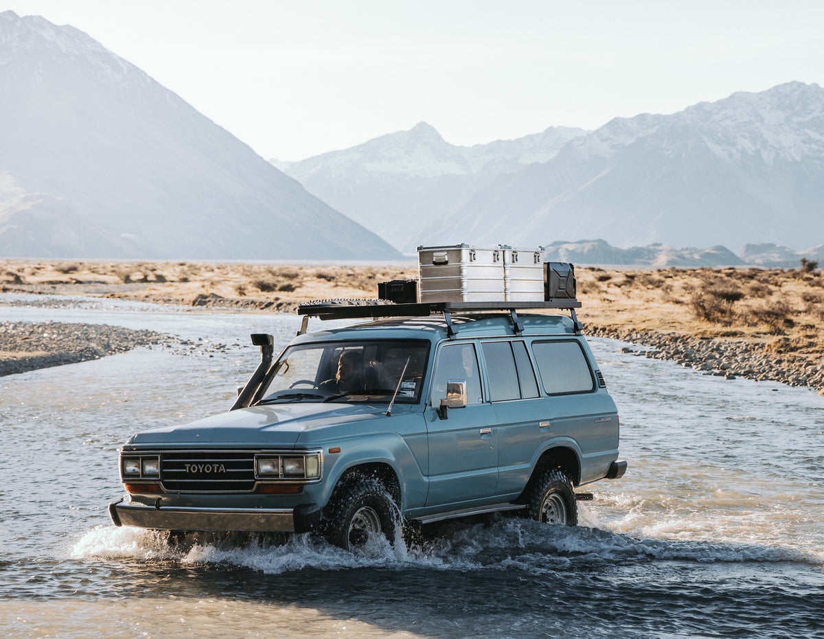 Toyota SUV fords a shallow river below snowy mountains with ZARGES K470-40839 cases on the roof rack