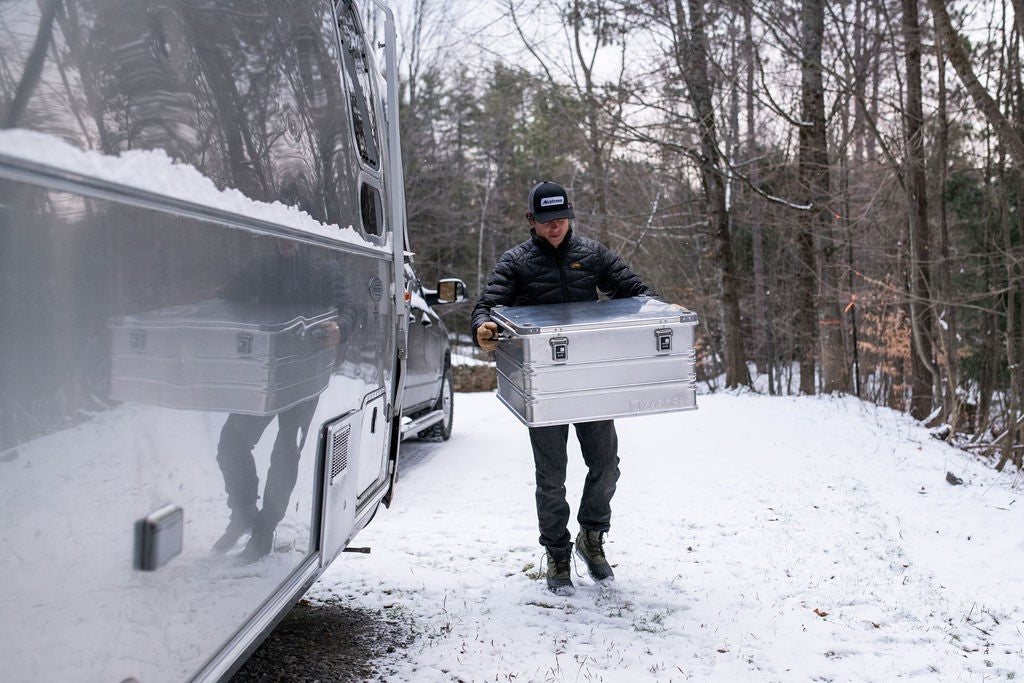 Man carrying a ZARGES K470-40842 aluminum case down a snowy driveway past a silver travel trailer