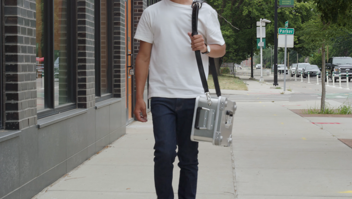Man in a white T-shirt walking a city sidewalk with an aluminum case on the ZARGES Leather Shoulder Strap