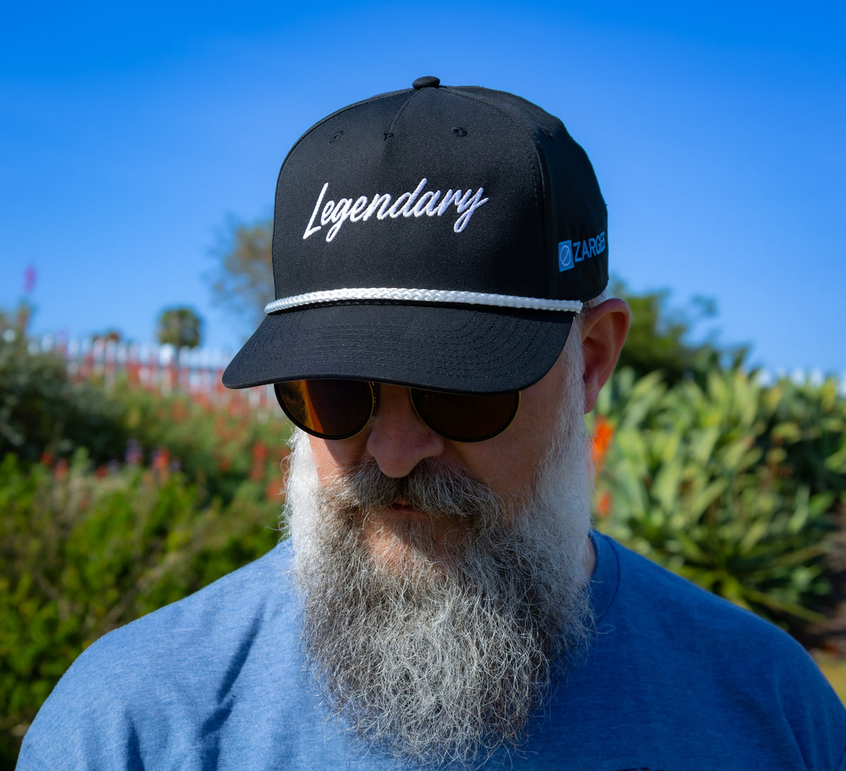 Bearded man in sunglasses wearing the ZARGES Legendary Black Rope Hat, front view under a blue sky
