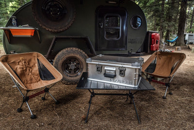 ZARGES K470-40568 case on a folding camp table between two chairs beside a teardrop trailer