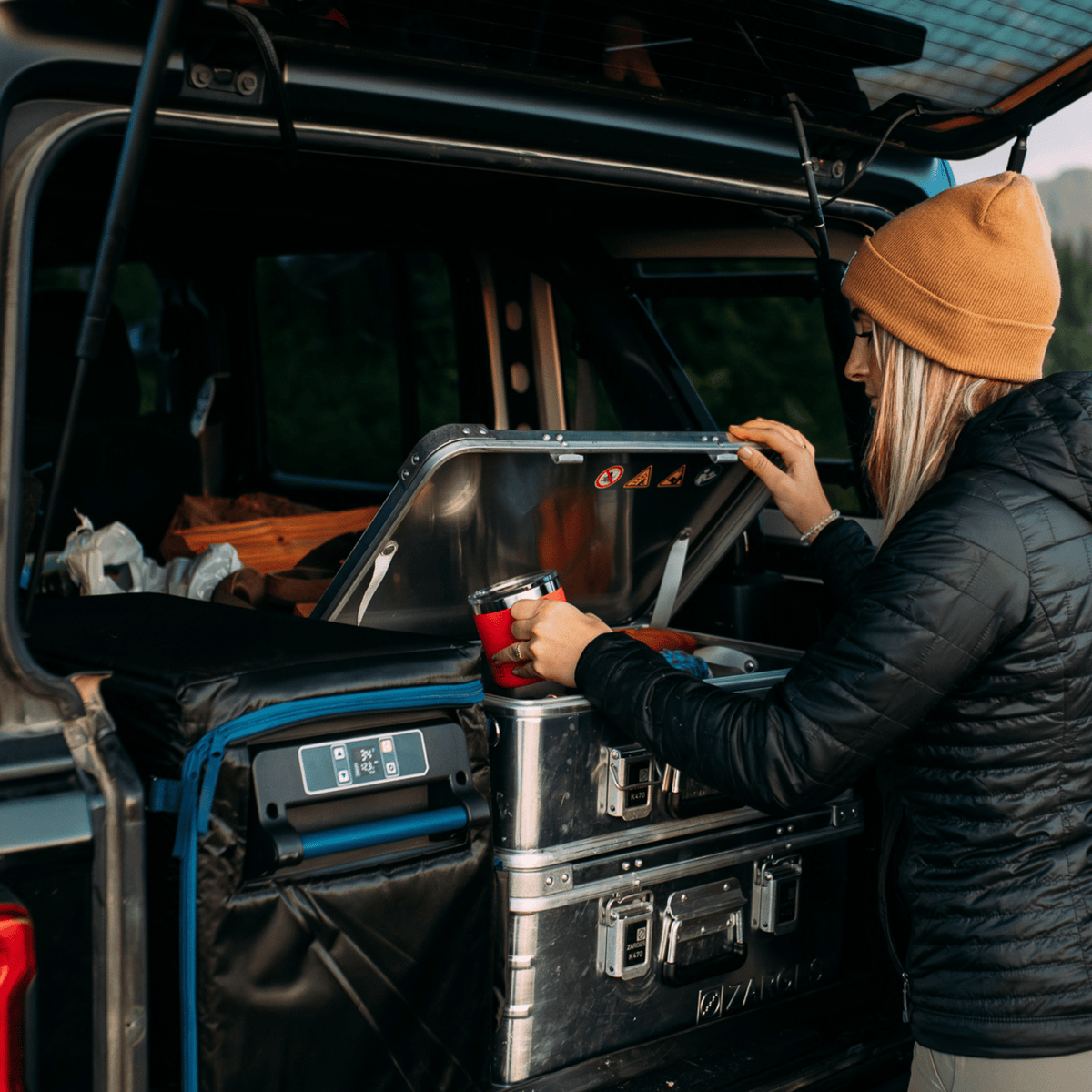 Woman in a beanie takes a mug from a stacked ZARGES K470-40810 case in the back of a vehicle
