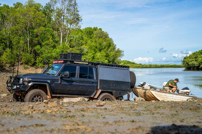 Off-road truck with a ZARGES K470-Matte Black-31013205 case on its roof rack by a boat at a muddy riverbank
