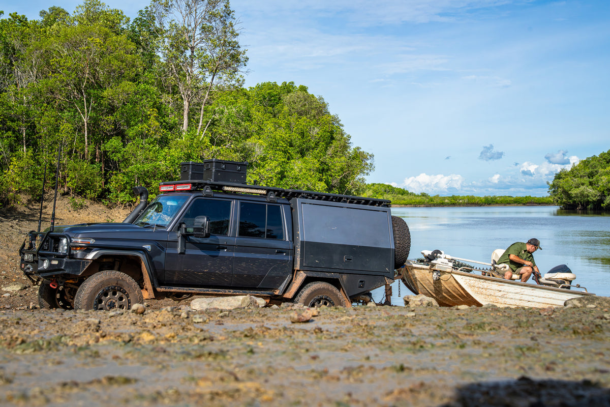 Off-road truck with a ZARGES K470-Matte Black-31013205 case on its roof rack by a boat at a muddy riverbank