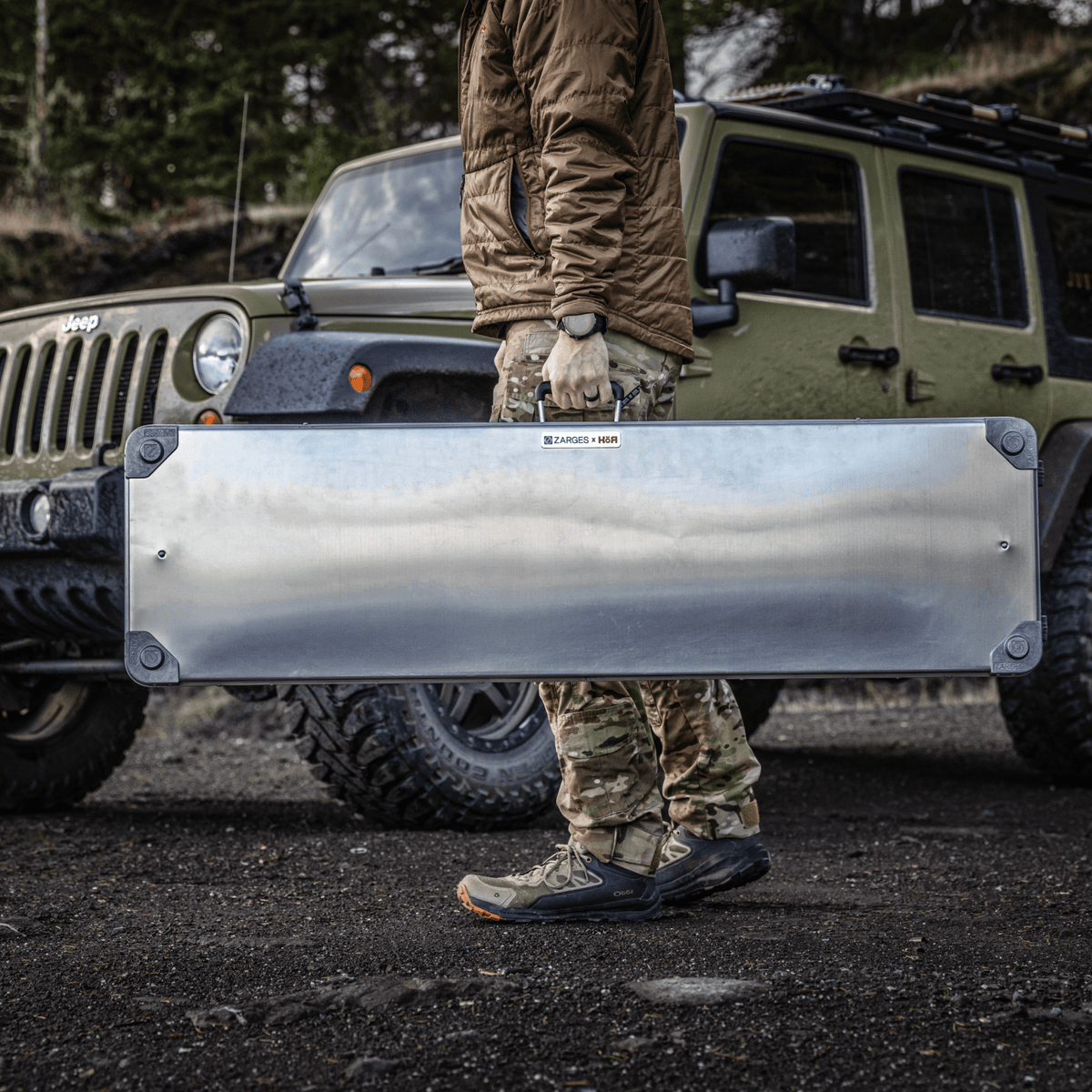 Person in camo carries a ZARGES Single Long Rifle KOR Kit case past a green Jeep
