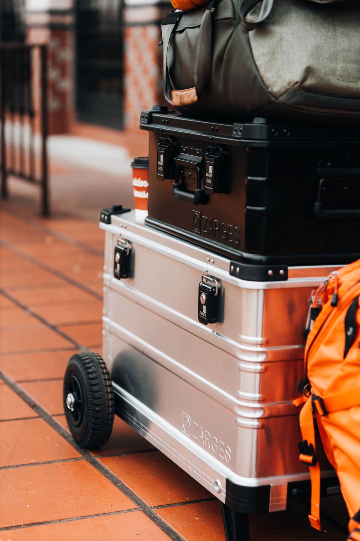 ZARGES K424 XC-41812 wheeled case carrying a black ZARGES case and duffel on a tiled sidewalk