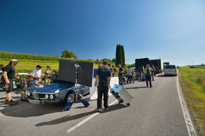 Film crew on a country road with a vintage blue car, one crew member rolling a ZARGES K424 XC-41813 case