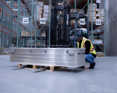 Warehouse worker crouching beside a long ZARGES K470-31004150 aluminum case on a pallet by a forklift
