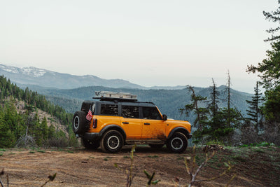 Orange SUV carrying a ZARGES K470-31011686 case on its roof rack, parked on a ridge above mountain valleys