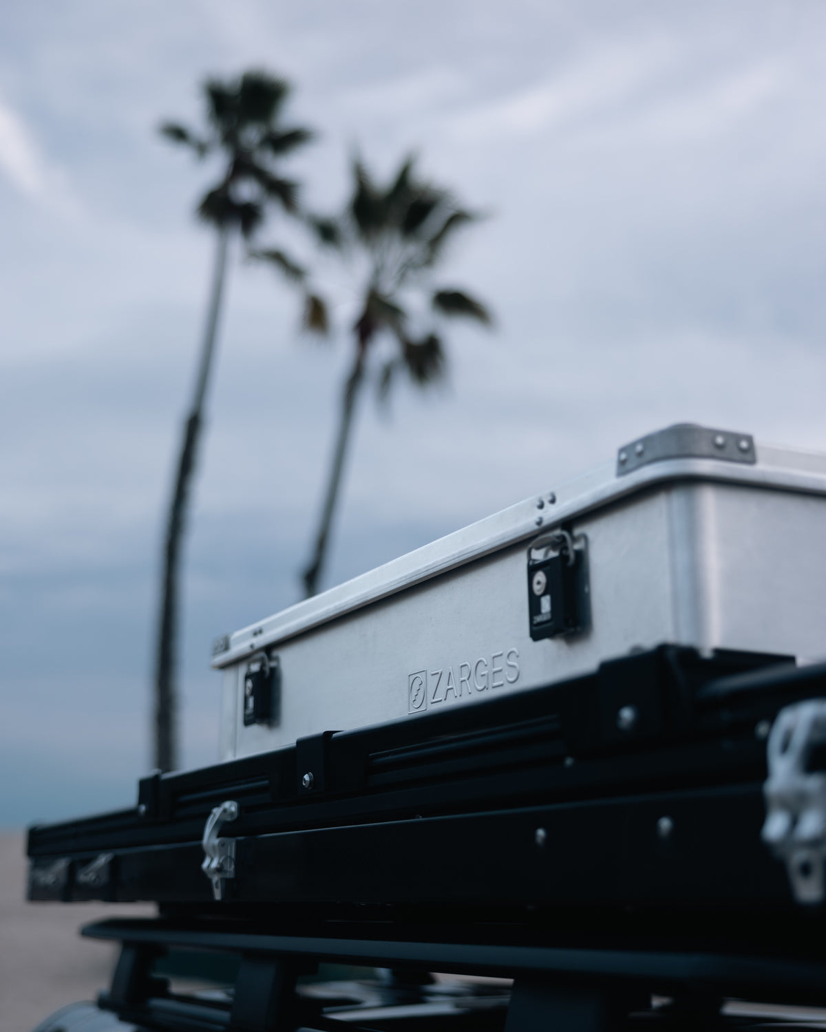 Close-up of a ZARGES K470-380031 case mounted on a vehicle roof rack under palm trees