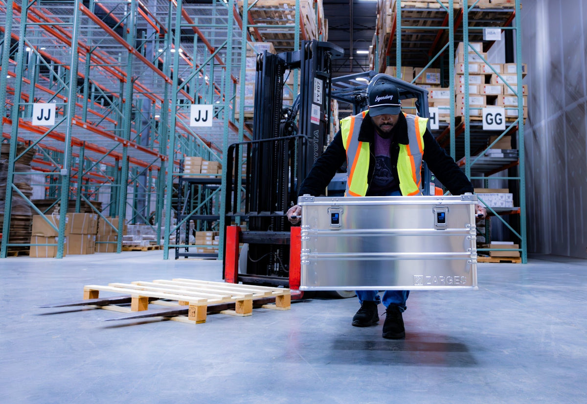 Warehouse worker in a safety vest carries a ZARGES K470-40565 case past a forklift and empty pallet