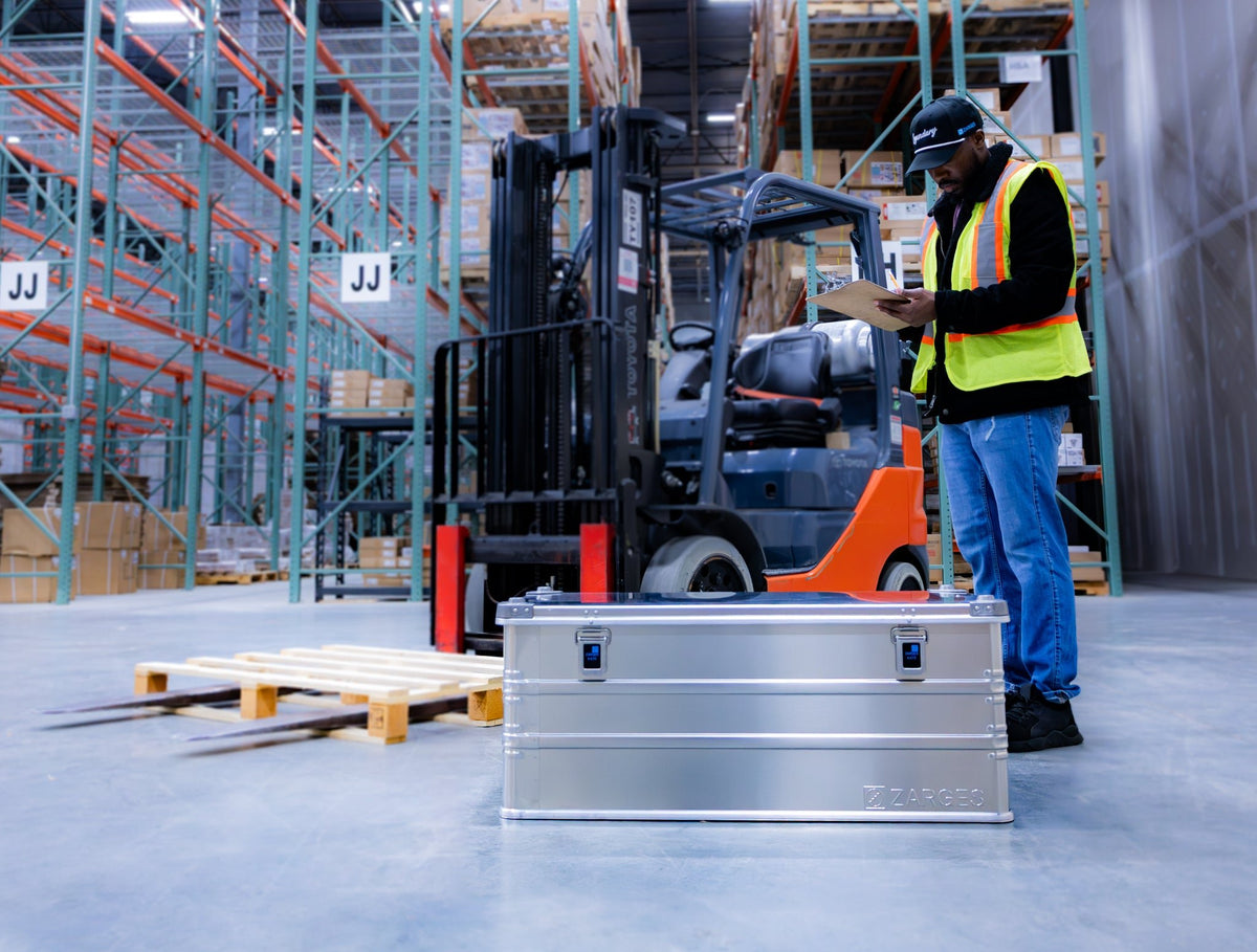 Warehouse worker with clipboard standing behind a ZARGES K470-40567 case beside a forklift