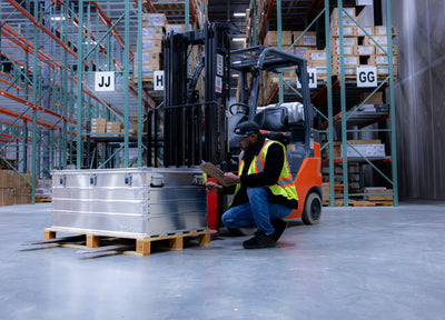 Worker kneeling beside a ZARGES K470-40580 case on a wooden pallet in a warehouse aisle