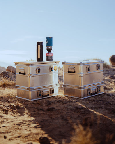 Two stacks of ZARGES K470-40678 cases on desert ground at sunset, a camp stove and bottle on top