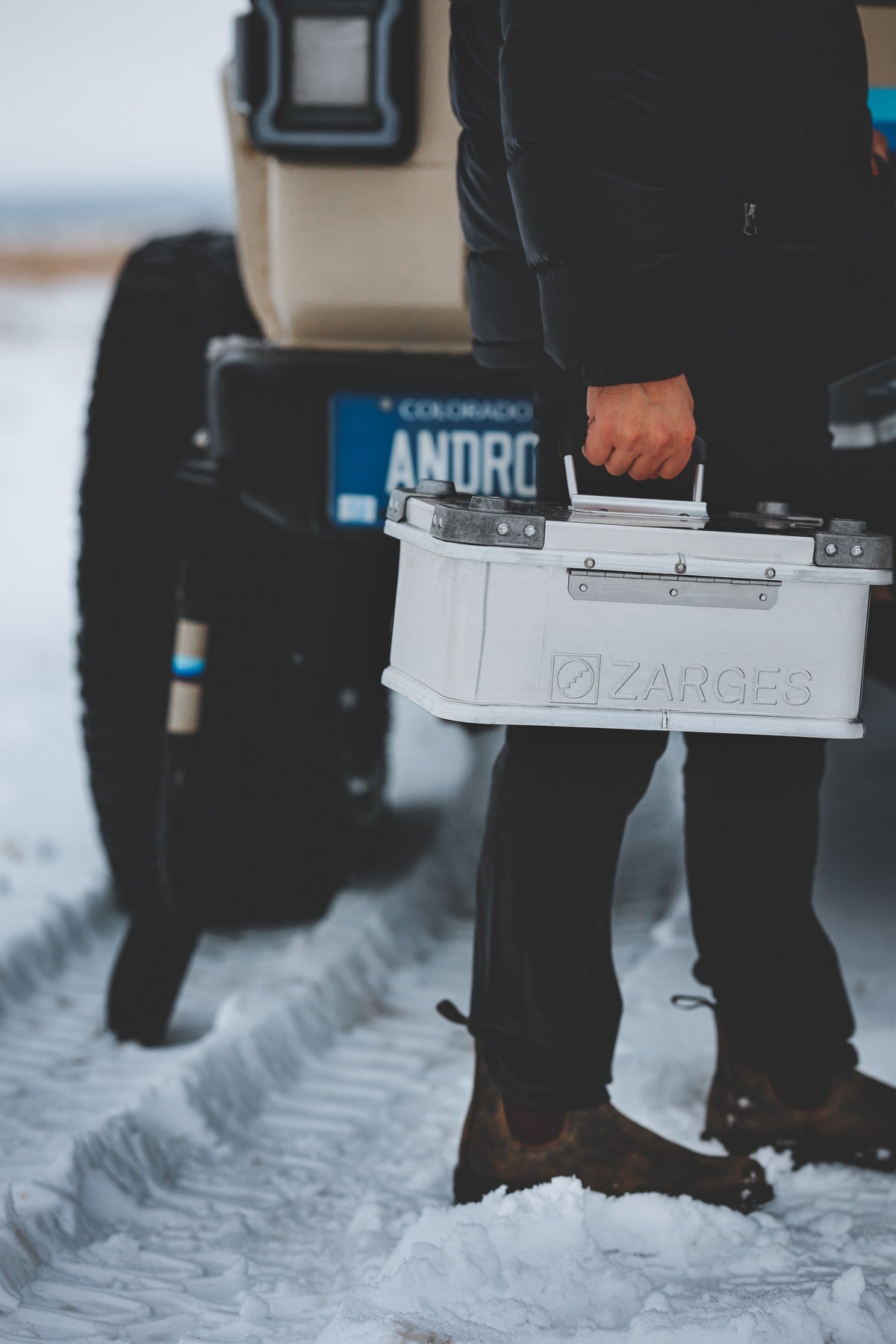 Person in boots carrying a white ZARGES K470-40835 case by its lid handle past an SUV in the snow