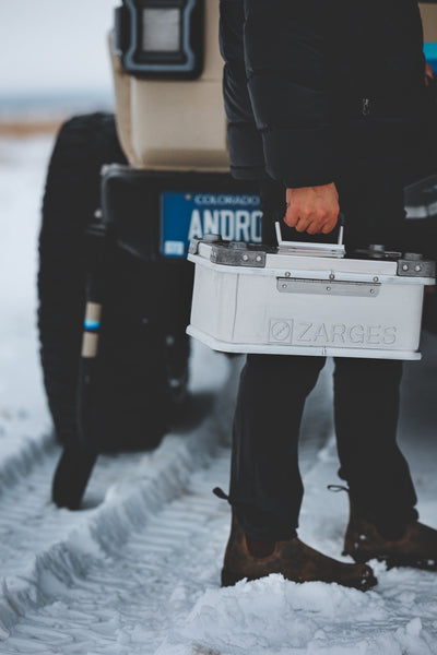 Person in boots carrying a white ZARGES K470-40835 case by its lid handle past an SUV in the snow