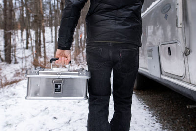 Person carrying a ZARGES K470-40835 aluminum case through a snowy forest beside a silver camper trailer