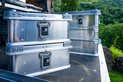Two ZARGES K470-40835 cases stacked on a truck bed with a larger ZARGES case behind, green hills beyond