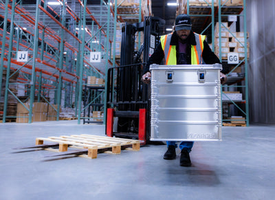 Warehouse worker in a hi-vis vest carrying a ZARGES K470-40836 aluminum case past a forklift