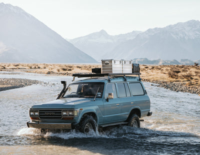 Toyota SUV fords a shallow river below snowy mountains with ZARGES K470-40839 cases on the roof rack