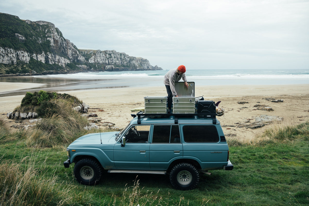 Man in an orange beanie opens a ZARGES K470-40839 case on the roof rack of a blue SUV at the beach