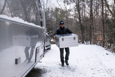 Man carrying a ZARGES K470-40842 aluminum case down a snowy driveway past a silver travel trailer