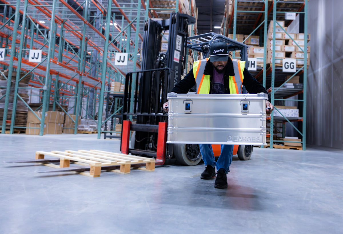 Warehouse worker in a hi-vis vest carrying a ZARGES K470-40842 aluminum case past a forklift