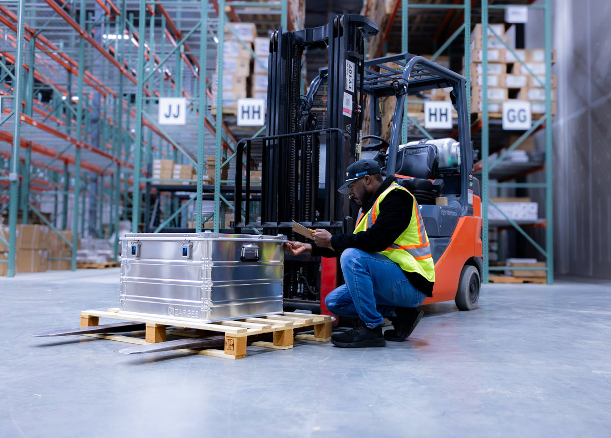 Warehouse worker kneeling with a clipboard beside a ZARGES K470-40843 case on a pallet