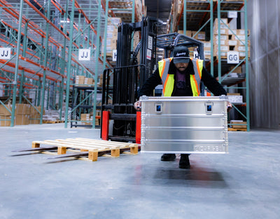 Warehouse worker in a hi-vis vest lifting a ZARGES K470-40843 aluminum case past a forklift