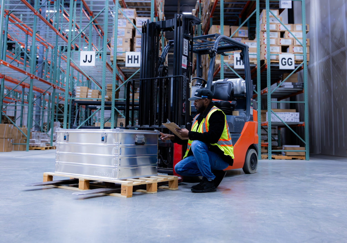Worker kneels with a clipboard beside a ZARGES K470-40846 case on a pallet by a forklift