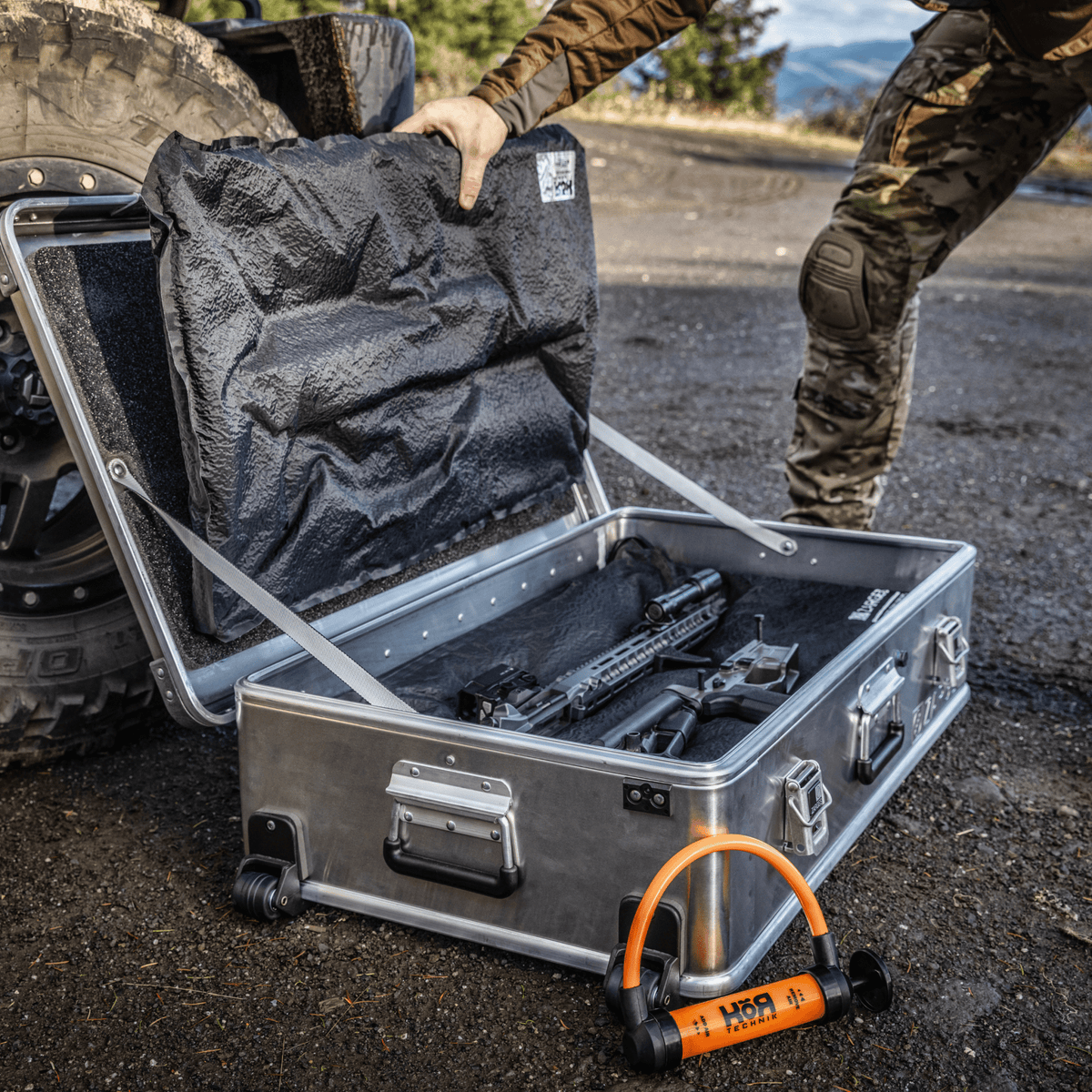 Hunter opens a wheeled ZARGES case beside a truck tire, KOR Takedown Kit panels over a broken-down rifle