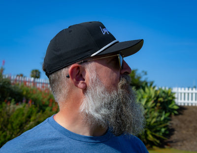 Bearded man wearing the ZARGES Legendary Black Rope Hat in profile, garden and white picket fence behind